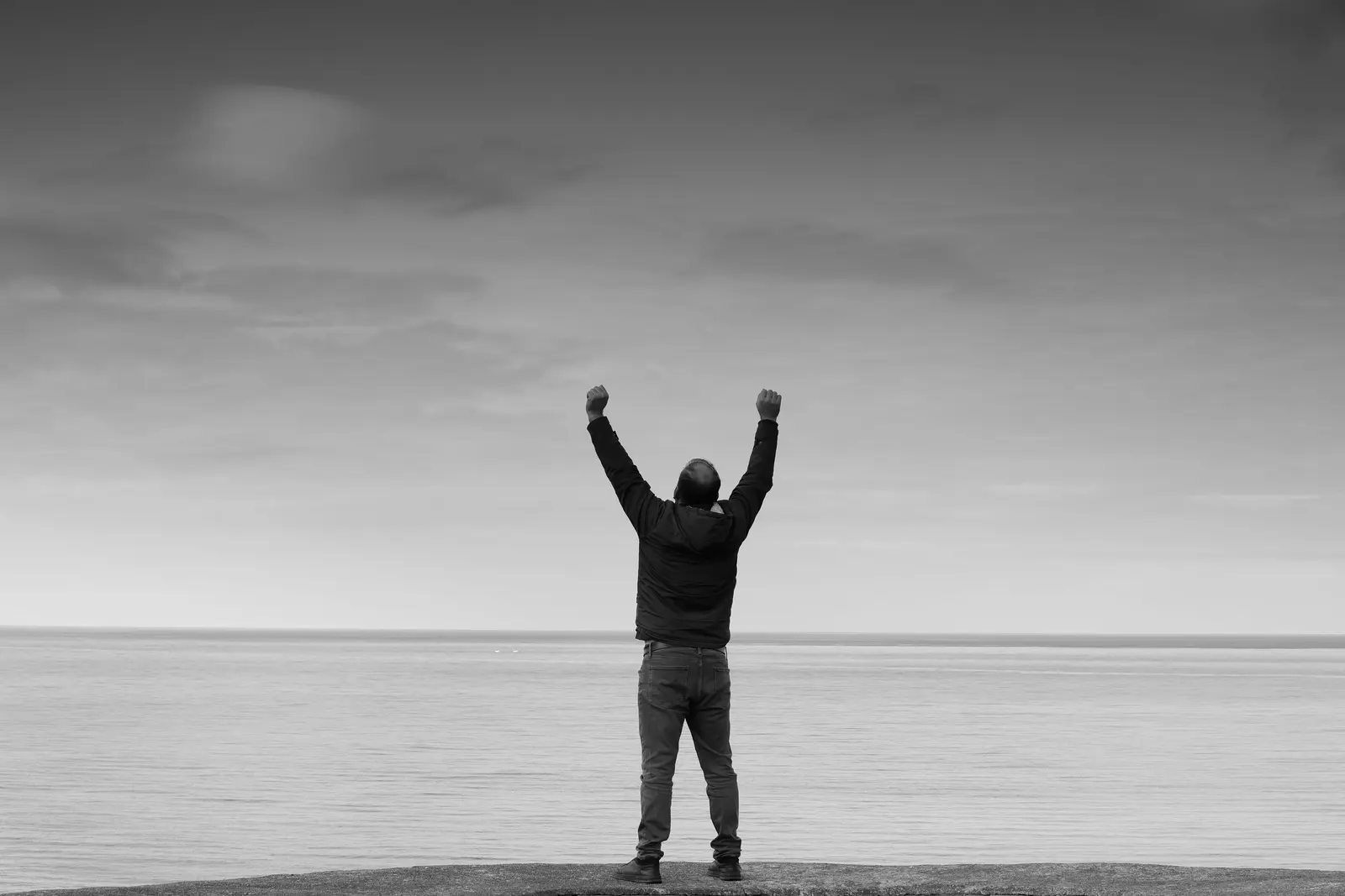 A man standing alone at the ocean, arms raised after working through a hard stretch